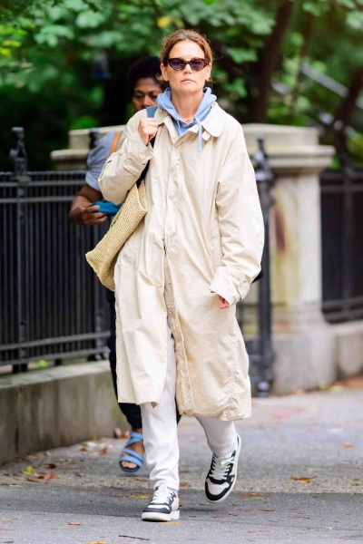 Look of the Day for August 8, 2024 features Katie Holmes in a khaki raincoat, blue hoodie, white sweatpants, and chunky basketball sneakers while out in New York City.